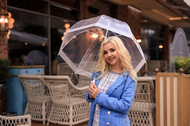 Retrato de joven y bella mujer rubia sosteniendo un paraguas transparente bajo la lluvia en una calle de la ciudad