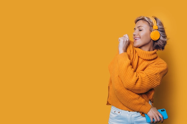 Retrato de una joven y bella mujer rubia vestida de amarillo bailando aislado en la pared amarilla escuchando meditando con auriculares amarillos.