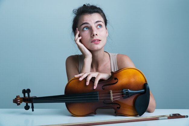 Retrato de una joven y bella mujer sentada a la mesa con un violín aislado sobre fondo azul.