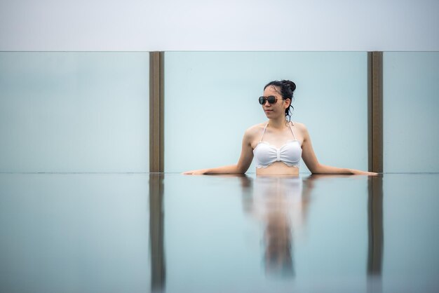 Retrato de una joven y bella mujer sonriente vistiendo traje de baño blanco y gafas mientras disfruta y se relaja en una piscina