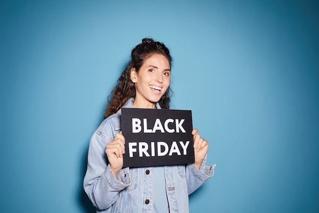 Retrato de joven bella mujer sosteniendo el cartel del Viernes Negro y sonriendo contra el fondo azul.