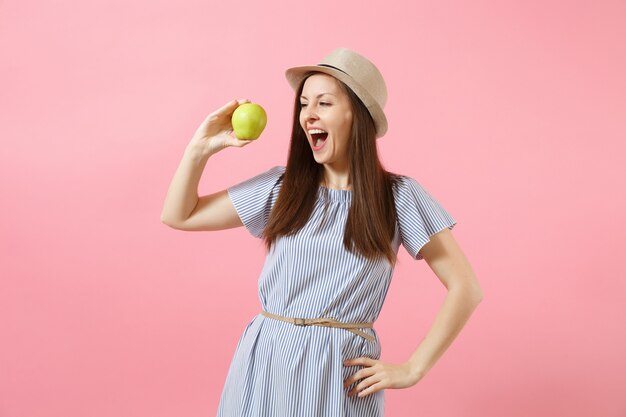 Retrato de joven bella mujer en vestido azul, sombrero de paja de verano sosteniendo, comiendo manzana verde fresca aislada sobre fondo rosa. Estilo de vida saludable, personas, concepto de emociones sinceras. Copie el espacio.