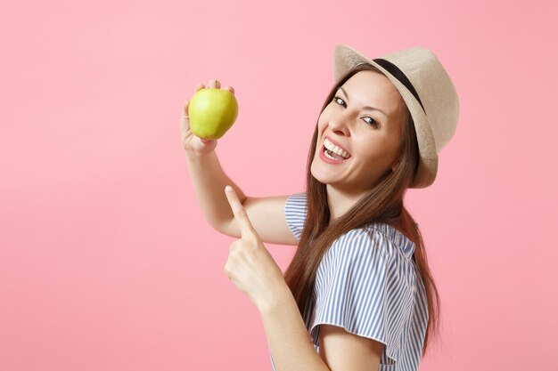 Retrato de joven bella mujer en vestido azul, sombrero de paja de verano sosteniendo, comiendo manzana verde fresca aislada sobre fondo rosa. Estilo de vida saludable, personas, concepto de emociones sinceras. Copie el espacio.