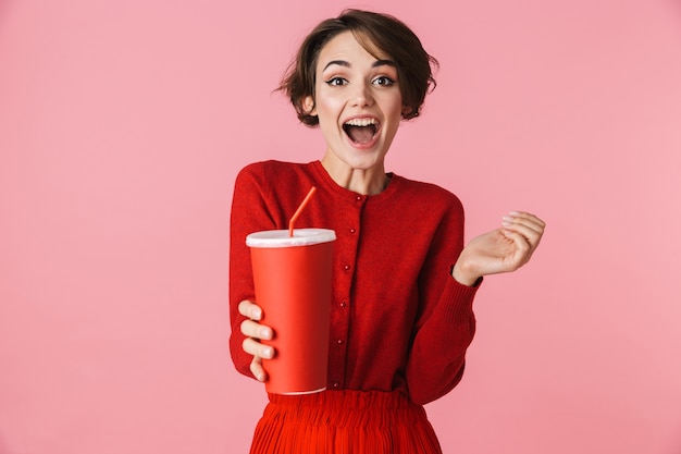 Retrato de una joven y bella mujer con vestido rojo que se encuentran aisladas sobre fondo rosa, sosteniendo un vaso de plástico con bebida