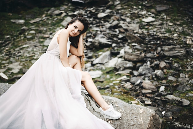 Retrato de una joven y bella novia caucásica con cabello largo mirando a la cámara mientras está sentado en una roca en las montañas con la cabeza apoyada en la mano sonriendo.