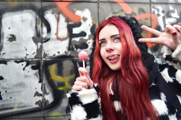 Retrato de una joven belleza adolescente en una sudadera con capucha con una deliciosa piruleta rosa, mostrando el signo de la paz en la cámara