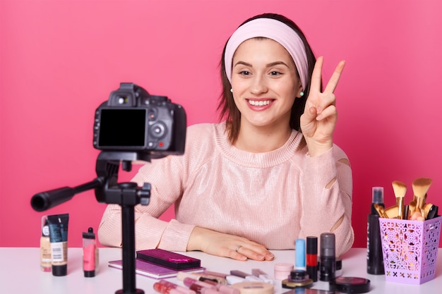 Retrato de joven blogger morena tomando video a través de cámara digital y mostrando el signo de la paz.