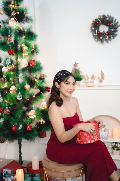 Retrato de una joven bonita y acogedora sentada sosteniendo un regalo de Navidad sonriendo vistiendo un vestido rojo en un salón de Navidad decorado en el interior