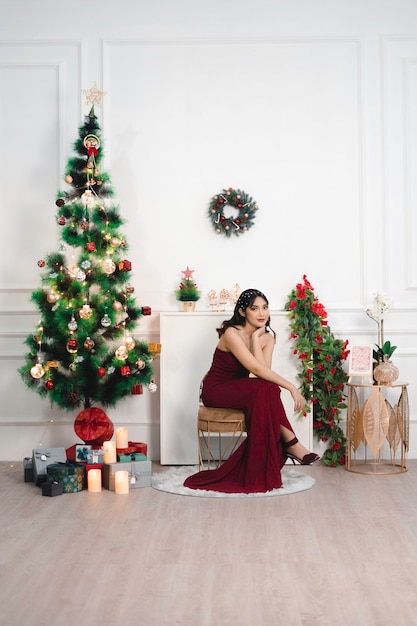 Foto retrato de una joven bonita y acogedora, siéntese sonriendo y use un vestido rojo en la sala de estar decorada de navidad en el interior