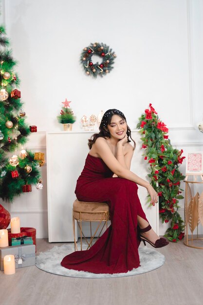 Retrato de una joven bonita y acogedora, siéntese sonriendo y use un vestido rojo en la sala de estar decorada de Navidad en el interior