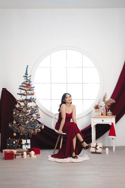 Retrato de una joven bonita y acogedora, siéntese sonriendo y use un vestido rojo en la sala de estar decorada de Navidad en el interior