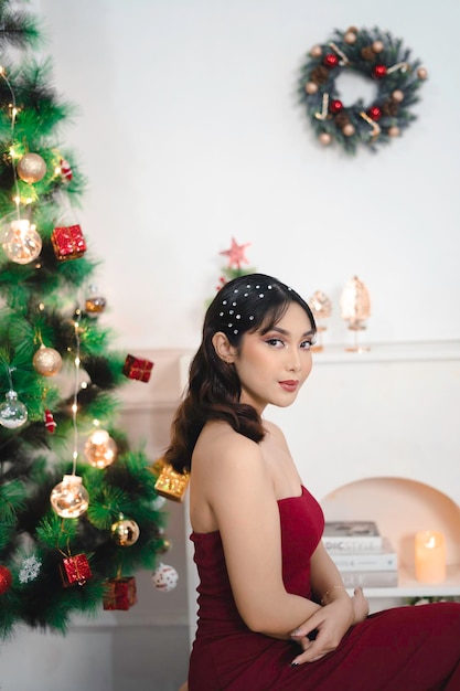 Retrato de una joven bonita y acogedora, siéntese sonriendo y use un vestido rojo en la sala de estar decorada de Navidad en el interior