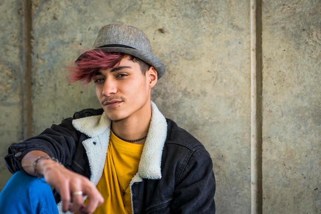 Retrato de un joven con el cabello púrpura al aire libre atractivo chico de moda sentado en el suelo con sombrero y peinado moderno adolescente de 18 años posando al aire libre