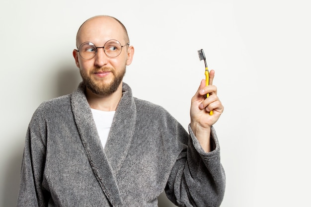 Retrato de un joven calvo con barba en bata y gafas y una cara sorprendida y sostiene un cepillo de dientes en la mano