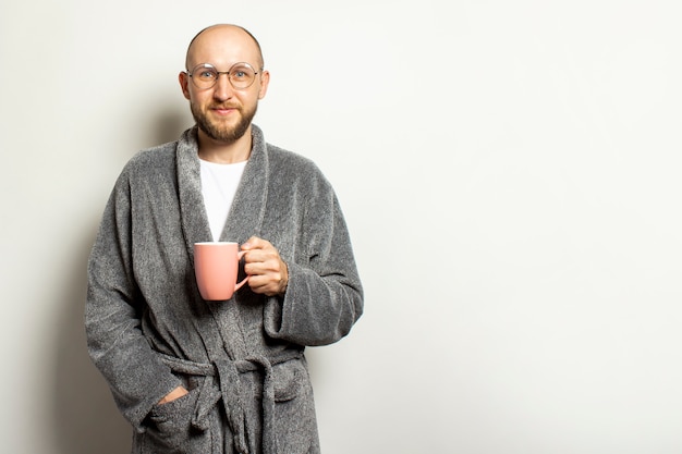 Retrato de un joven calvo con barba en bata y gafas sosteniendo una taza de café caliente