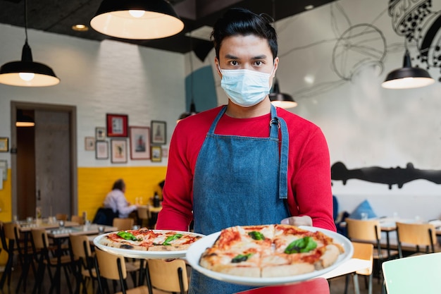 Retrato de un joven camarero asiático con mascarilla sirviendo pizzas