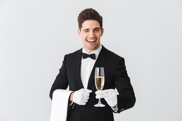 Foto retrato de un joven camarero sonriente en esmoquin le ofrece una copa de champán sobre la pared blanca