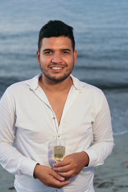 Retrato de joven caminando con una copa de vino blanco por la orilla del mar.