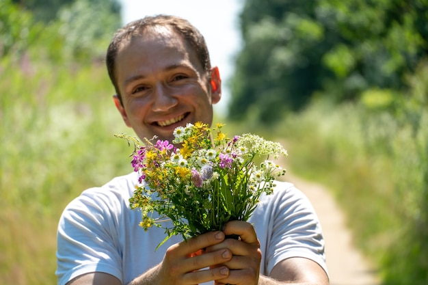 Retrato de un joven con una camiseta blanca y un ramo de flores en las manos Un chico posa en el fondo de un campo de flores y un camino de tierra en el pueblo La cara de una persona feliz