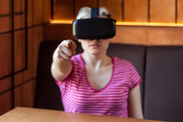 Retrato de una joven con una camiseta rosa sentada en un café, usando auriculares de realidad virtual y viendo videos, jugando en un simulador y señalando con el dedo a la cámara. interior, estilo de vida