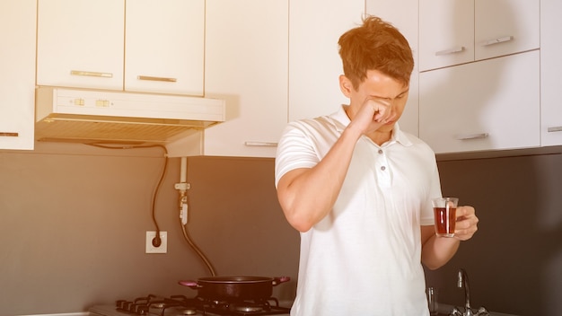 Retrato de joven cansado y soñoliento está bebiendo un té por la mañana en la cocina de casa. Bosteza y se frota los ojos, tratando de despertar. Temprano en la mañana de un día laborable.