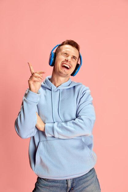 Retrato de un joven con capucha escuchando música con auriculares y cantando emocionalmente aislado sobre fondo rosa Concepto de emociones música