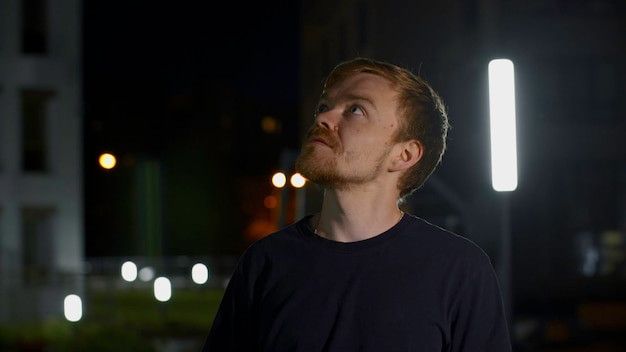 Retrato de un joven con capucha de pie al aire libre en la ciudad por la noche y posando para los medios de comunicación de la cámara