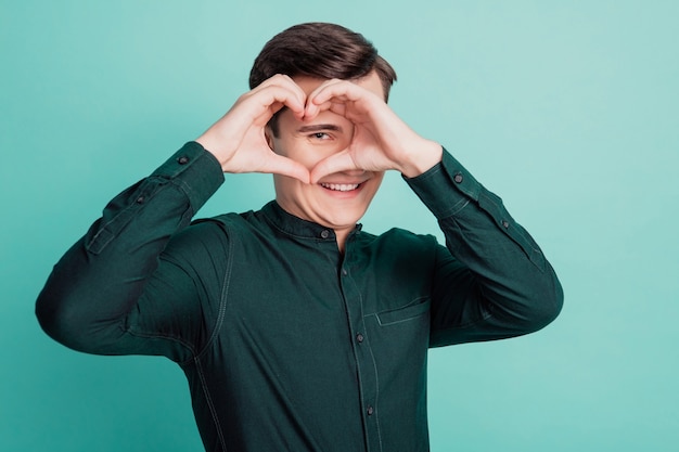 Retrato de joven cariñoso mostrar el símbolo del corazón dedos cubren el ojo sobre fondo verde azulado