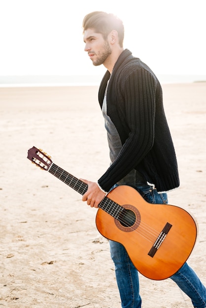 Retrato de un joven casual caminando por la orilla del mar con guitarra