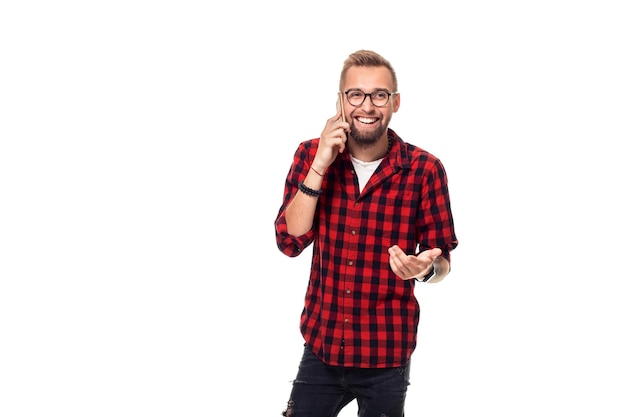 Retrato de un joven casual hablando por teléfono y sonriendo mientras mira hacia otro lado, en algún lugar. Disparo de estudio. Joven hipster con camisa a cuadros y gafas con fondo blanco