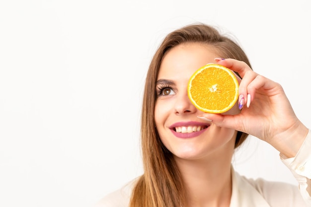 Retrato de una joven caucásica alegre que cubre los ojos con una rodaja de naranja con una camisa blanca sobre un fondo blanco