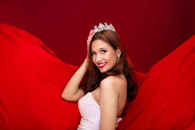 Retrato de una joven caucásica delgada que lleva un vestido largo de noche con lentejuelas blancas, una chica hermosa posa medio cuerpo sintiendo emoción sonrisa feliz, tela roja que agita el fondo