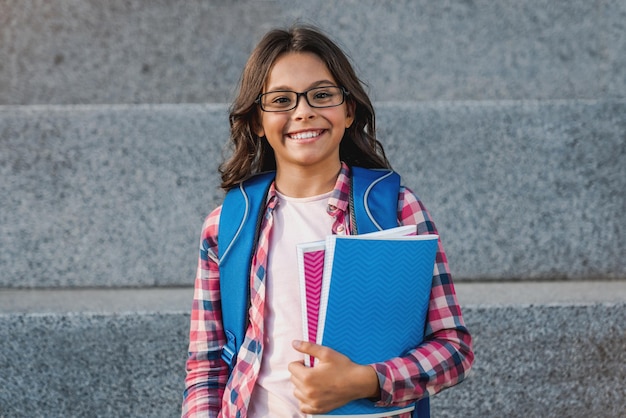Retrato de una joven caucásica feliz con mochila sosteniendo un cuaderno fuera de la escuela primaria