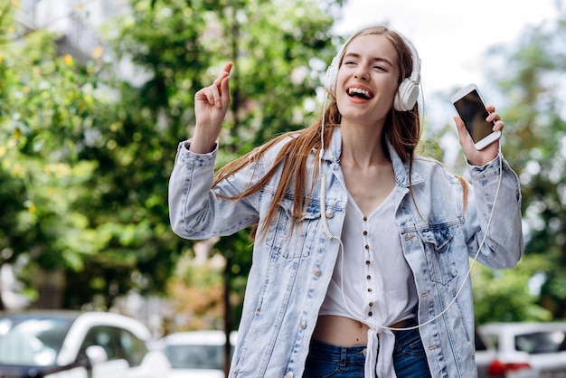 Retrato de la joven caucásica que se siente encantada mientras usa un teléfono inteligente y auriculares en la ciudad Niña bailando en la calle de verano