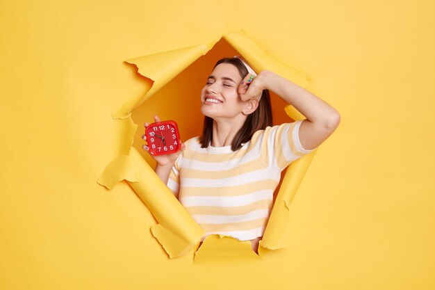 Retrato de una joven caucásica satisfecha con camisa a rayas y banda para el cabello posando en un agujero de papel amarillo de pie y estirando el cuerpo después de dormir se despierta de buen humor sosteniendo un despertador