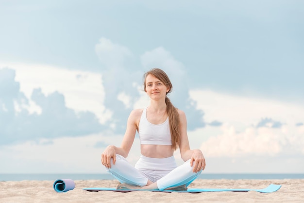 Retrato de una joven caucásica sentada en una alfombra de yoga en el fondo del mar Practica yoga al aire libre en la orilla del mar