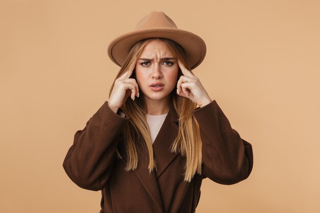 Retrato de joven caucásica con sombrero y abrigo pensando y tocando sus sienes aisladas en beige