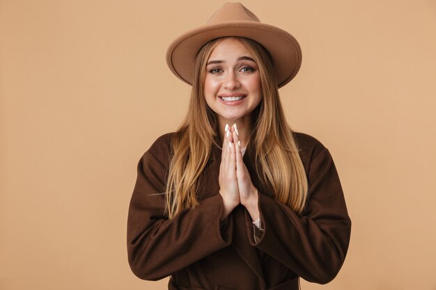 Retrato de joven caucásica con sombrero riendo y manteniendo las palmas juntas aisladas en beige