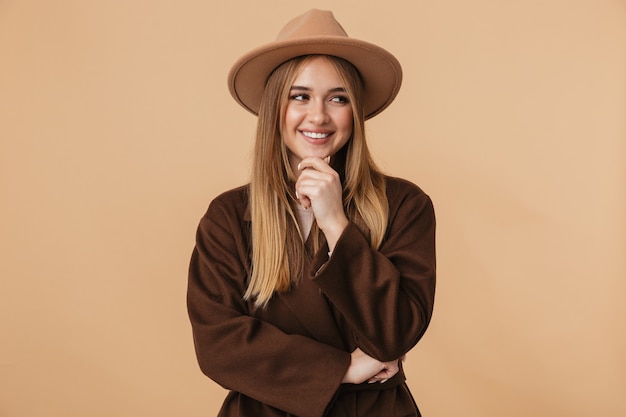 Retrato de joven caucásica con sombrero riendo y tocando su barbilla aislado en beige