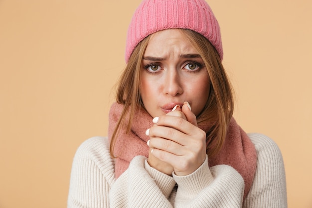 Retrato de joven caucásica vistiendo gorro y bufanda de invierno temblando y sintiendo frío aislado en beige