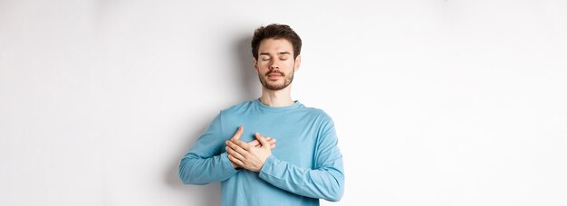 Retrato de joven caucásico con barba de pie con las manos en el corazón y los ojos cerrados soñando abo