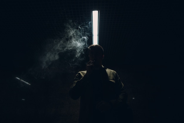 Retrato de joven caucásico fumar cigarrillo electrónico en la oscuridad con luz de línea.