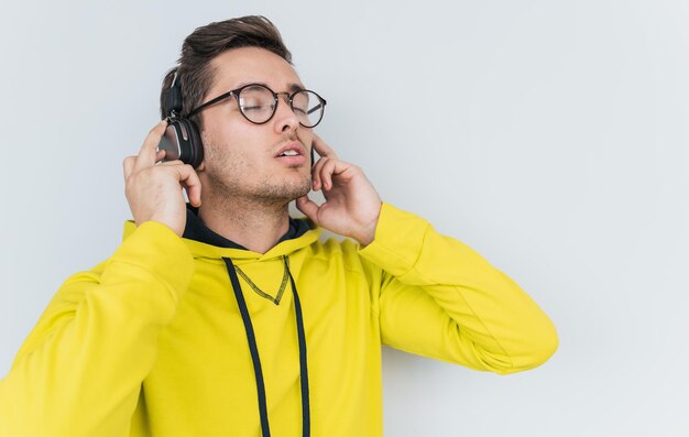 Retrato de un joven caucásico relajado vestido con una sudadera con capucha amarilla deportiva sosteniendo auriculares con ambas manos escuchando música favorita con los ojos cerrados aislados en el espacio de copia de fondo blanco para el texto