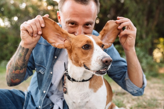 Retrato de joven caucásico en ropa de mezclilla jugando con su perro canino en el parque