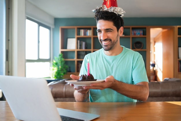 Retrato de joven celebrando un cumpleaños en una videollamada desde casa con un portátil y un pastel. Nuevo concepto de estilo de vida normal.