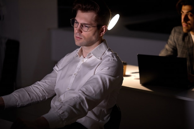 Retrato de joven centrado cansado en anteojos trabajando de forma remota a altas horas de la noche en la oficina oscura usando la computadora, pensando en la fecha límite, en ropa formal. Exceso de trabajo, pensamientos negativos, concepto de fecha límite