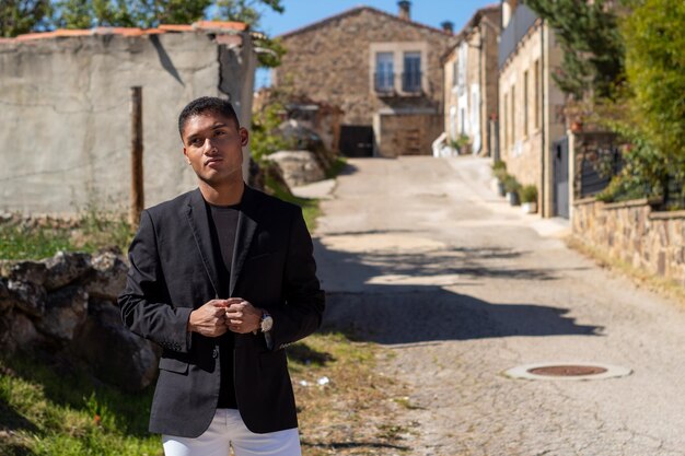 retrato, de, un, joven, en, chaqueta negra, y, pantalón blanco, con, un, rural, vivienda, área, atrás, él