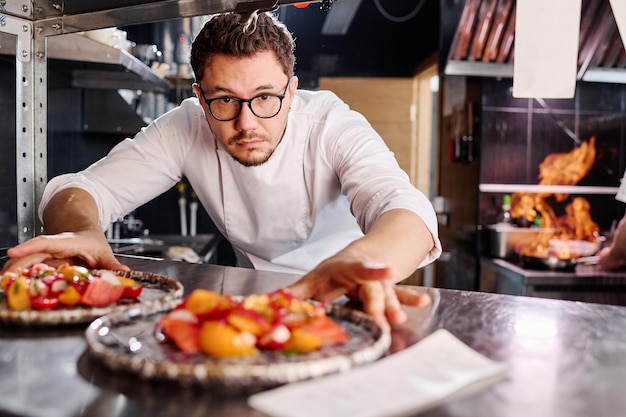 Retrato de joven chef en anteojos poniendo platos de ensalada en el mostrador de la cocina para la entrega