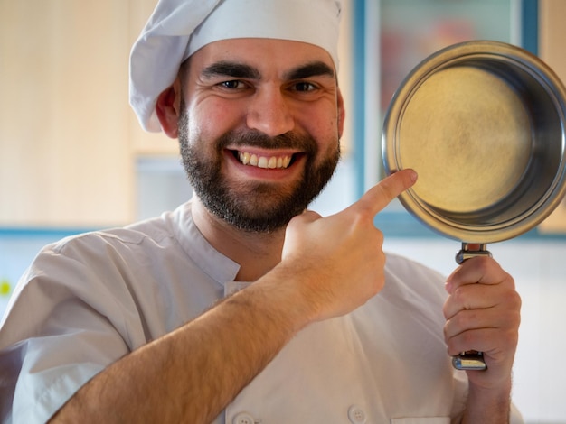 Foto retrato de un joven chef barbudo sonriendo mientras señala una cacerola inoxidable mirando a la cámara