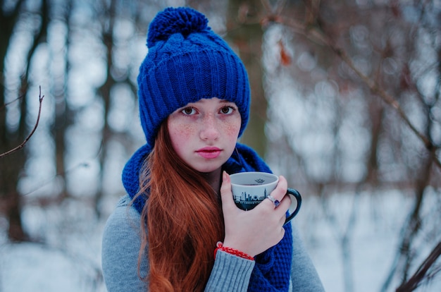 Retrato de joven chica de pelo rojo con pecas en azul gorro de lana tejida y bufanda con taza de té en día de invierno.